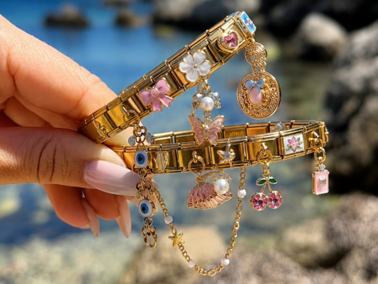 Two gold Italian charm bracelets held up at the water, hung with a butterfly, cherries, a bow, a shell and an evil eye
